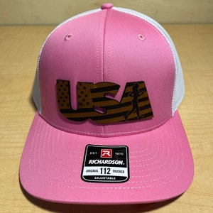 Puede incluir: Gorra de camionero rosa y blanca con un gráfico de EE. UU. marrón que presenta una figura corriendo y estrellas. La gorra tiene una correa ajustable con el logotipo de Richardson y el texto "Original 112 Trucker Adjustable".