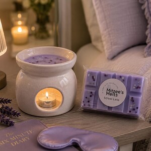 May include: A white ceramic wax warmer with a lit tea light candle, a rectangular bar of purple wax melts labeled Mitzee's Melts Stress Less, a purple sleep mask, and a book titled The Six Minute Diary, all arranged on a wooden surface.