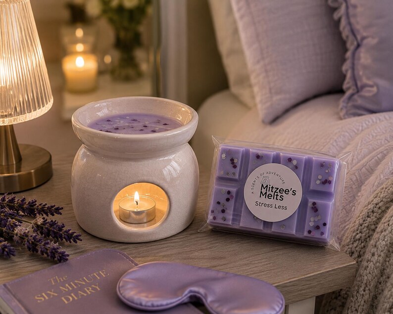 May include: A white ceramic wax warmer with a lit tea light candle, a rectangular bar of purple wax melts labeled Mitzee's Melts Stress Less, a purple sleep mask, and a book titled The Six Minute Diary, all arranged on a wooden surface.