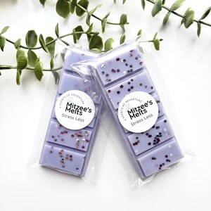 May include: Two rectangular light purple wax melt bars packaged in clear plastic sleeves. Each bar features a white circular label with the text Mitzee's Melts Stress Less and a small candle icon. The wax is embedded with small dark glitter flecks.