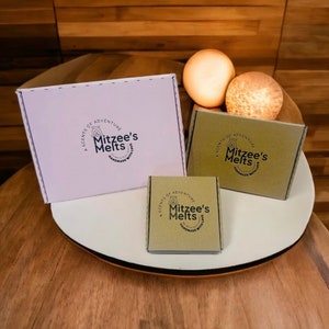 May include: Three rectangular cardboard boxes of varying sizes featuring the Mitzee's Melts logo. One large white box and two smaller brown boxes are displayed, each printed with the text A Scents of Adventure Mitzee's Melts Handmade with Love.