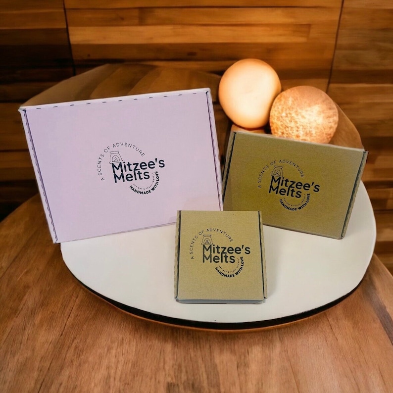 May include: Three rectangular cardboard boxes of varying sizes featuring the Mitzee's Melts logo. One large white box and two smaller brown boxes are displayed, each printed with the text A Scents of Adventure Mitzee's Melts Handmade with Love.