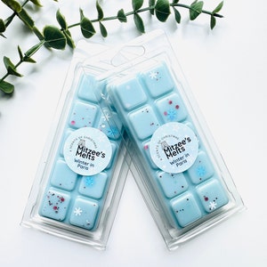 May include: Two clear plastic packs of wax melts, each containing six individual squares. The wax melts are light blue with white snowflakes and pink glitter. The packs have a round sticker with the text "A Scents of Christmas Mitzee's Melts Winter in Paris".