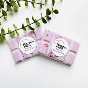 May include: Two rectangular light purple wax melts packaged in clear plastic. Each bar features a circular white label with the text A SCENTS OF ADVENTURE Mitzee's Melts Dreams Unstoppable. The wax is embedded with small pink and dark glitter flecks.