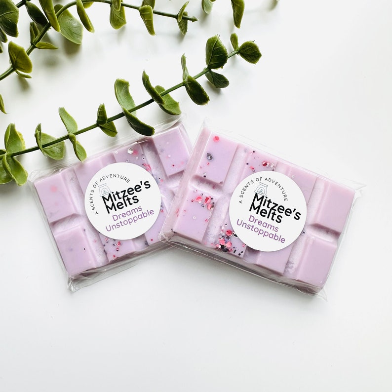 May include: Two rectangular light purple wax melts packaged in clear plastic. Each bar features a circular white label with the text A SCENTS OF ADVENTURE Mitzee's Melts Dreams Unstoppable. The wax is embedded with small pink and dark glitter flecks.