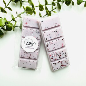 May include: Two rectangular light purple wax melts featuring a segmented bar design with embedded red, black, and silver glitter particles. One bar is inside a clear plastic sleeve with a circular white label reading Mitzee's Melts Dreams Unstoppable.