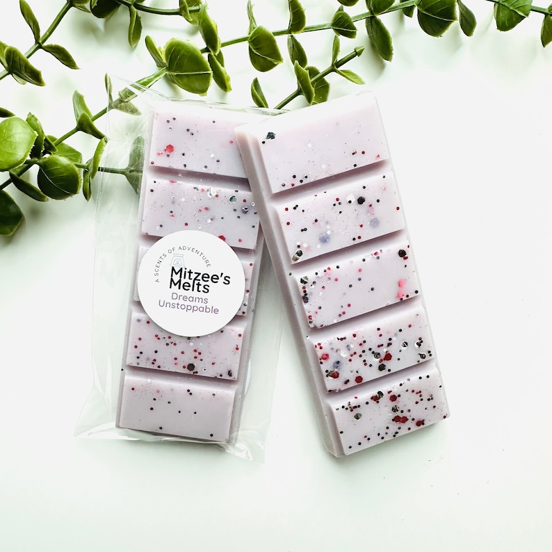 May include: Two rectangular light purple wax melts featuring a segmented bar design with embedded red, black, and silver glitter particles. One bar is inside a clear plastic sleeve with a circular white label reading Mitzee's Melts Dreams Unstoppable.