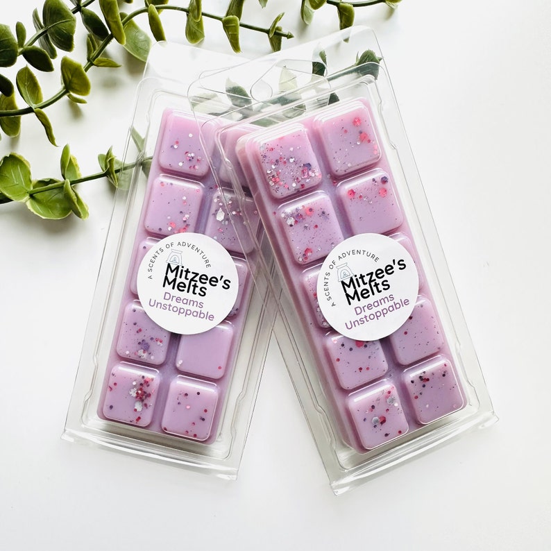 May include: Two clear plastic clamshell packages containing light purple wax melts with embedded pink, silver, and purple glitter. Each package features a circular white label with the text Mitzee's Melts and Dreams Unstoppable.