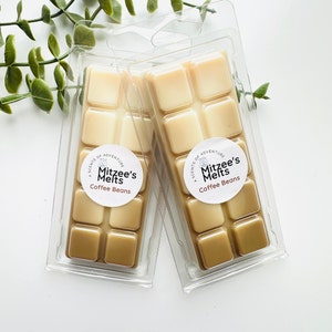 May include: Two clear plastic clamshells each containing eight square wax melts. The wax cubes are two-toned, with lighter and darker shades of brown. Each package has a round label that reads "Mitzee's Melts Coffee Beans."
