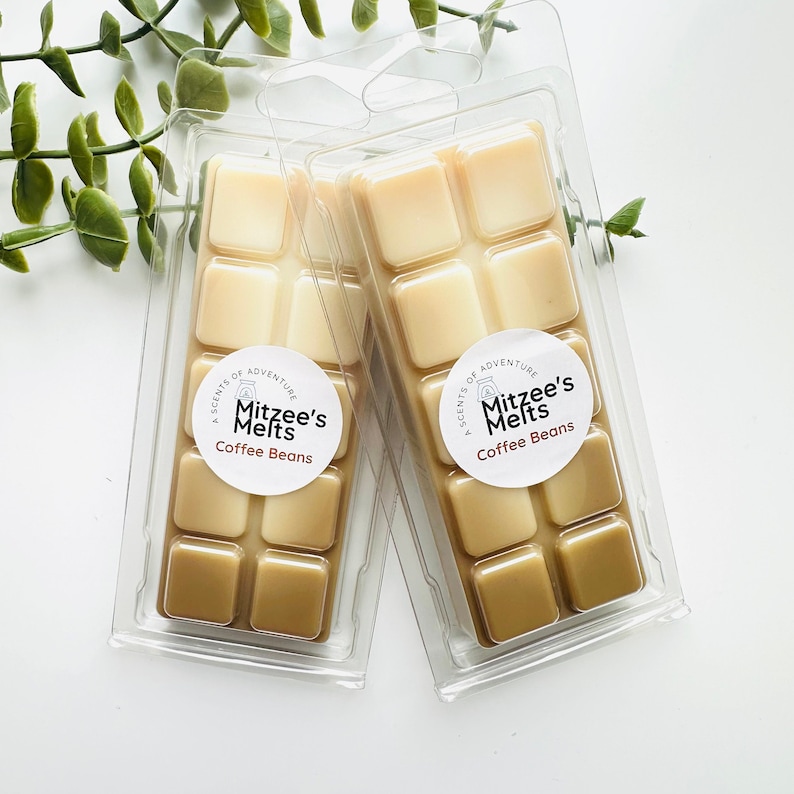May include: Two clear plastic clamshells each containing eight square wax melts. The wax cubes are two-toned, with lighter and darker shades of brown. Each package has a round label that reads "Mitzee's Melts Coffee Beans."