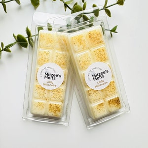 May include: Two clear plastic containers of wax melts, each with six squares of scented wax. The wax is a light yellow colour with gold glitter. The labels on the containers read "A Scent of Adventure, Mitzee's Melts, Lady Millionaire".