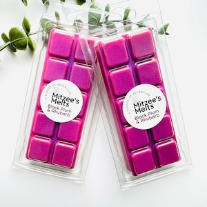 May include: Two rectangular clear plastic clamshell packages containing ten square magenta wax melts each. A circular white label on each package features the text Mitzee's Melts, A Scents of Adventure, and Black Plum & Rhubarb in black font.