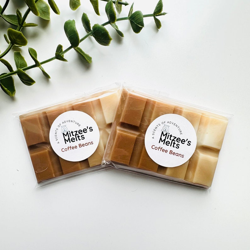 May include: Two wax melt bars in clear packaging, each with a coffee bean scent. The bars are divided into rectangular sections, with shades of brown and tan. Each package has a white circular label with the text "Mitzee's Melts Coffee Beans". Green leaves are in the background.