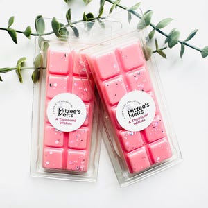 May include: Two pink wax melts in clear plastic packaging. The wax melts are square and have a glittery finish. The packaging has a white label with the text "A Scents of Adventure Mitzee's Melts A Thousand Wishes".