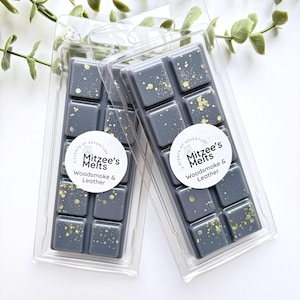 May include: Two rectangular wax melt bars in clear plastic packaging. Each dark grey bar features ten square segments topped with gold glitter. A circular white label on each reads: A Scents of Adventure, Mitzee's Melts, Woodsmoke & Leather.