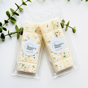 May include: Two rectangular plastic clamshell packages containing off-white wax melts. Each wax melt bar is divided into ten segments embedded with multicoloured glitter. A circular label reads A Scents of Christmas Mitzee's Melts Jingle Bell.