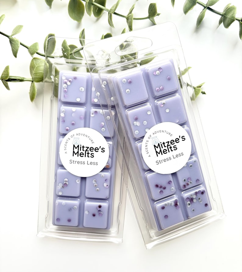 May include: Two rectangular clear plastic clamshell packages containing light purple wax melts. Each wax melt bar is divided into ten square segments and topped with small white and purple glitter. A circular white label reads Mitzee's Melts Stress Less.