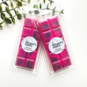 May include: Two packs of pink wax melts with a speckled design. Each pack contains eight square melts and a label that reads "Mitzee's Melts Festive Cranberry". The wax melts are in clear plastic packaging.