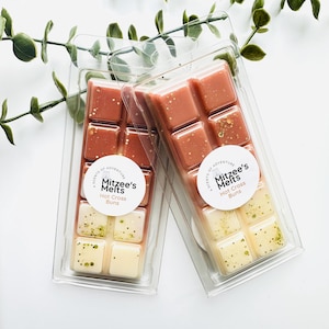 May include: Two rectangular plastic clamshell packages containing wax melts. Each package features a label reading Mitzee's Melts Hot Cross Buns. The wax is divided into ten segments, with the top half in a dusty rose colour and the bottom half in cream, both accented with small gold and green glitter flecks.