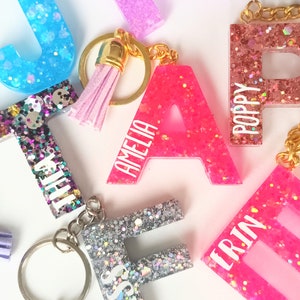 May include: A set of colourful glitter resin letter keychains with the names Amelia, Poppy, Erin, and Issy. Each letter is attached to a gold chain and a tassel.
