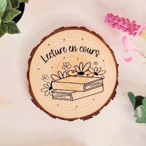 Puede incluir: Un posavasos redondo de madera con el texto "Lecture en cours" y un dibujo de dos libros con flores en la parte superior. El posavasos está sobre una superficie blanca.