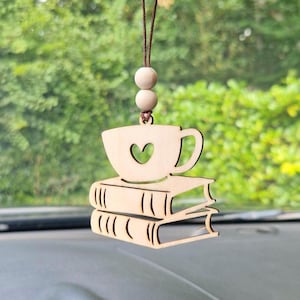 May include: A wooden car charm featuring a coffee cup with a heart cutout, resting on two stacked books. The charm is suspended by a brown string with two wooden beads. The background is a blurred view of green foliage.