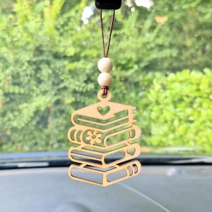 Peut inclure: Un charm de voiture en bois représentant une pile de livres avec des découpes en forme de cœur et de fleur. Le charme est suspendu par une ficelle marron et deux perles en bois. Fond vert et feuillu.