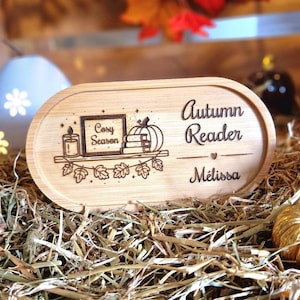 Peut inclure: Panneau ovale en bois avec les mots "Autumn Reader" et "Melissa" gravés. Il présente une étagère avec une bougie, des livres et une citrouille. Les mots "Cosy Season" sont également gravés.