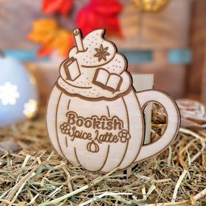Puede incluir: Decoración de madera con forma de calabaza con la inscripción "Bookish Spice Latte". El diseño incluye crema batida, una pajita y libros abiertos. El fondo está borroso con decoraciones otoñales.