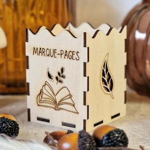 Peut inclure: Un porte-marque-pages en bois avec le texte "MARQUE-PAGES" et un motif de livre ouvert et de feuilles. Le porte-marque-pages est en bois clair et a un design découpé.