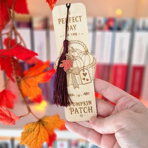 Può includere: Un segnalibro in legno chiaro con un'illustrazione incisa al laser di un cesto pieno di zucche. Il testo recita "PERFECT DAY IN THE PUMPKIN PATCH". Una nappa color bordeaux è fissata in alto, decorata con un piccolo ciondolo a foglia d'acero.