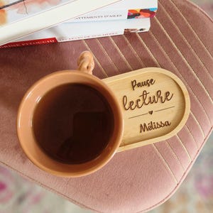 以下が含まれることがあります： 紅茶が入った桃色のマグカップが、「Pause lecture」と「M&eacute;lissa」と刻まれた木製のコースターの上に置かれています。ピンク色のベルベットの表面に、本の山が背景にあります。