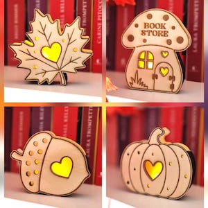 Op de afbeelding: Een verzameling van vier verlichte houten ornamenten in de vorm van een esdoornblad, een paddenstoelvormig gebouw met de tekst BOOK STORE, een eikel en een pompoen. Elk ornament heeft een hartvormige uitsparing met een geel licht.