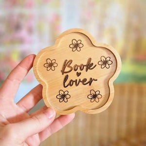Op de afbeelding: Een houten onderzetter in de vorm van een bloem met de woorden "Book lover" in het midden gegraveerd. Kleine bloemmotieven sieren de randen. De onderzetter is lichtbruin van kleur.