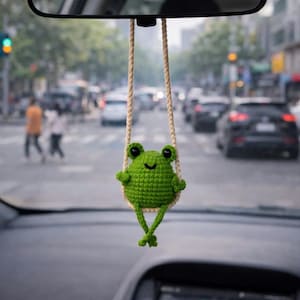 May include: A green crocheted frog car charm hanging from the rearview mirror. The frog has large black eyes and is seated on a swing made of beige rope. The background shows a city street with cars and pedestrians.