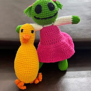 May include: A crocheted green doll with a pink dress and a yellow crocheted duck. The doll has a white crocheted shirt and green hair.