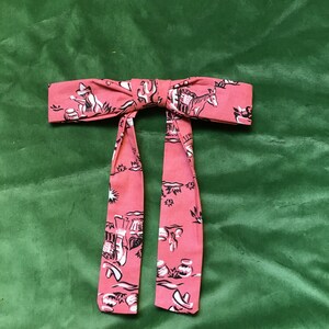 May include: A pink bow tie with a black and white print of a desert scene. The print includes cacti, a donkey, and a person riding a donkey.