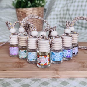May include: A collection of small glass car air freshener bottles with wooden caps and braided hanging cords. Each bottle features a unique label with text including Castle of Wishes, Riverside Orleans, Fiji Waves, Sunshine State, and Banshee Flight.