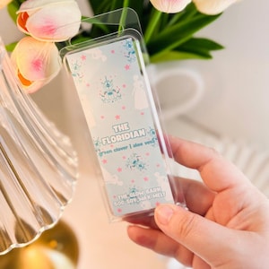 May include: A clear plastic clamshell package containing a soy wax melt. The label is light blue with a pattern of white silhouettes, blue chandeliers, and pink stars. Text reads: THE FLORIDIAN, green clover | aloe vera, THE GLOW BARN 500 SOY WAX MELT.