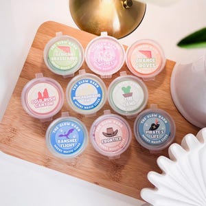May include: Nine small, round, clear plastic containers with lids, each featuring a label from The Glow Barn. Labels include African Grasslands, Castle of Wishes, Orange Groves, Cold Rush Canyon, Philharmagic Pie, The Land, Banshee Flight, Frontier, and Pirate's Life.