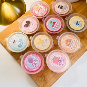 May include: Ten small, round, clear plastic containers filled with colourful wax melts, each topped with a circular label featuring The Glow Barn branding and unique scent names like Popcorn Cart, Coffee & Castles, Krispy Treats, Cherry Glacier, Be Our Guest, Ice Cream Parlour, Magical Waffles, Bayou Adventures, Candy Emporium, and Peach Whip.