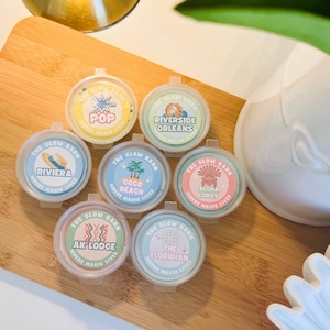 May include: Seven small, round, clear plastic containers with lids, each featuring a circular label with the text The Glow Barn Where Magic Lives. The labels display unique designs and names: Pop, Riverside Orleans, Riviera, Coco Beach, Aloha Lobby, Ak Lodge, and The Floridian.
