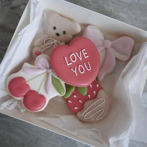 Peut inclure: Une boîte de biscuits décorés, dont un ours en peluche, un cœur avec le texte "LOVE YOU", une fraise trempée dans du chocolat, un nœud et un motif de cerise. Les biscuits sont roses, blancs et verts.