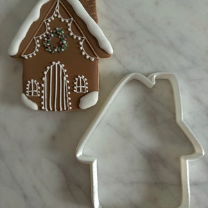 Könnte beinhalten: Ein Lebkuchenhaus-Keks, verziert mit weißem Zuckerguss und einem roten und grünen Kranz. Neben dem Keks liegt ein weißer Lebkuchenhaus-Ausstecher aus Kunststoff.
