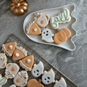 Könnte beinhalten: Eine Auswahl an herbstlich verzierten Keksen mit Zuckerguss in Form von Kürbissen, Geistern, Tassen mit Sahnehäubchen, Tortenstücken und dem in Schreibschrift geschriebenen Wort fall, angerichtet auf einem weißen, geisterförmigen Teller und einem Gitter.