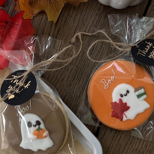 Peut inclure: Deux biscuits d'Halloween emballés individuellement. L'un est marron avec un fantôme tenant une citrouille, l'autre orange avec un fantôme et une feuille d'érable. Les deux ont "Boo!" écrit dessus. Chaque biscuit a une étiquette "Merci!".