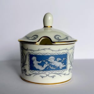 Puede incluir: Un azucarero de cerámica color crema con tapa a juego y borde dorado. El cuerpo presenta un panel rectangular azul con figuras en relieve blanco de dos querubines, rodeado de delicados adornos en gris y detalles en oro.
