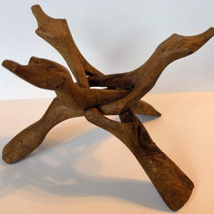 May include: A brown wooden display stand crafted from two interlocking pieces of carved wood. The stand features four legs and four upward-facing extensions, each shaped like a stylized animal head with visible eyes and a pointed snout.