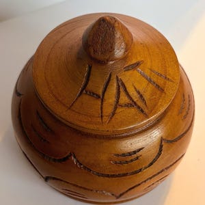 May include: A round, medium-brown wooden container with a matching lid. The lid features a pointed center knob and the hand-carved text "HAITI" across the top. The body of the container is decorated with carved, scalloped patterns around the sides.