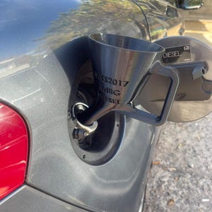 May include: A black funnel with a handle is inserted into a car's fuel tank. The funnel has the text "MKS2017 ItdBG DIESEL" printed on its side. The car is gray, and the fuel door is open.